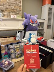 A hand holds a red "SUMMIT COLLECTIBLES" mystery box in front of a display featuring Pokémon trading cards and a purple Gengar plush toy.