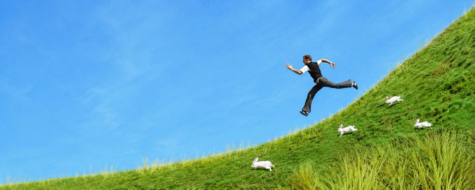 A surreal photo-manipulation image showing a man in business attire appearing to fall or fly across a green hillside under a blue sky, with small white rabbit figures scattered on the grass below.