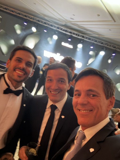 Three men in formal suits smile for a selfie at an indoor gathering, with a drink visible in the foreground and bright event lighting overhead.