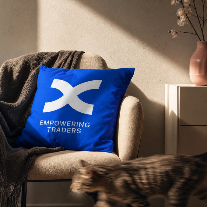 A cozy armchair is draped with a brown throw and features a bright blue pillow displaying a white "X" logo with the text "EMPOWERING TRADERS," visually illustrating the comfort implied by "the only thing comfier than a green candle." A vase with cherry blossoms sits on a side table nearby, and a cat darts across the foreground.
