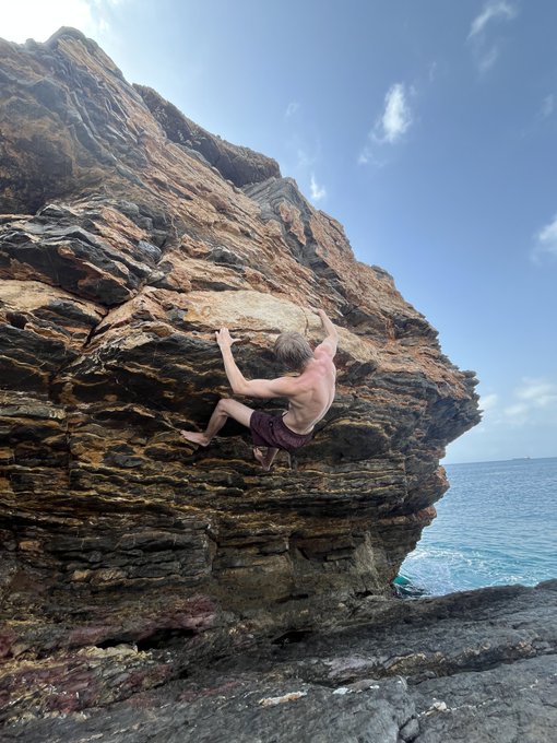 A shirtless man in red shorts bouldering on a large rocky coastal cliff overlooking the ocean under a partly cloudy sky.
