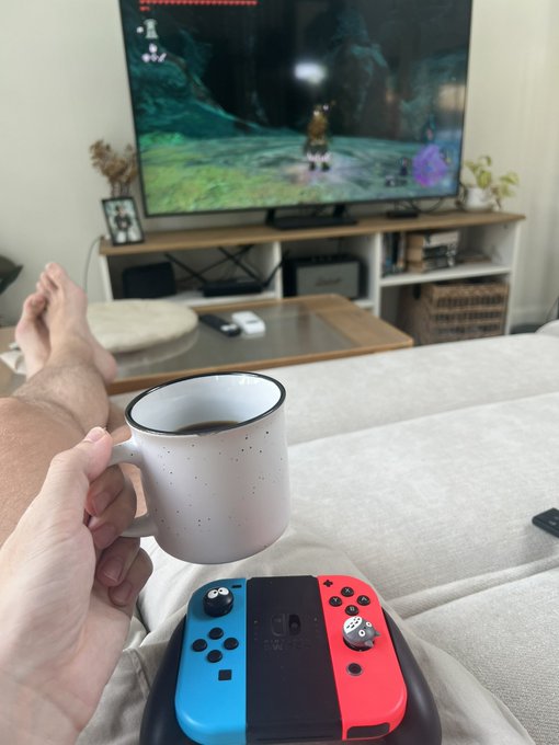 A first-person view of someone relaxing on a couch holding a mug of coffee, with a Nintendo Switch controller resting on their lap and a video game playing on a TV in the background.
