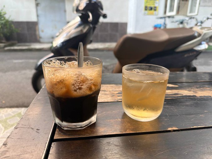 Two glasses of iced beverages—one dark coffee and one lighter tea—sit on a weathered wooden table outside, with motorbikes parked on the street in the background.