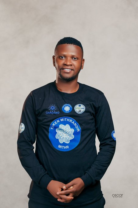The image features a man wearing a black long-sleeved shirt with blue designs on it. The man has short hair and is smiling at the camera while posing for the photo. The background of the image is white, which contrasts nicely with the man's dark clothing. There are no other objects or people in the picture besides the man and his surroundings.