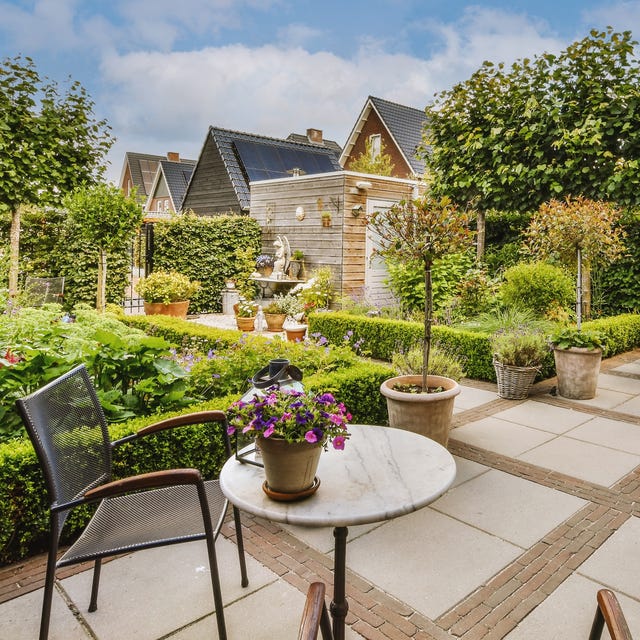 A photograph of a beautifully landscaped garden patio featuring a round marble table with purple petunias, a black mesh chair, neatly trimmed hedges, potted plants, and traditional houses in the background.