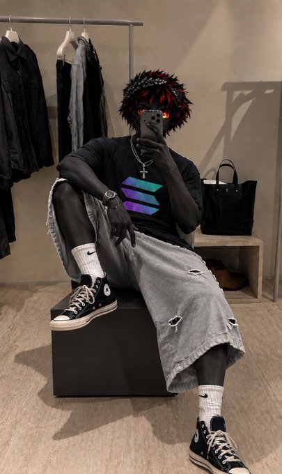 A person with dark skin and dark, dreadlocked hair with red highlights, wearing sunglasses, a black t-shirt with a colorful logo, ripped gray denim shorts, and black high-top Converse sneakers, taking a mirror selfie with a smartphone in a clothing store.