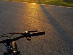 A first-person view of bicycle handlebars with the warm light of sunset reflecting off a wet paved road ahead.