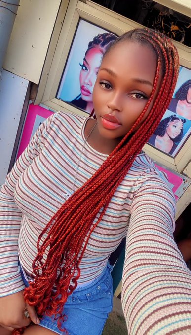 A selfie of a young woman with long red braided hair wearing a striped sweater and denim shorts, standing in front of a shop displaying photos of hairstyles.