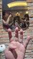 Low-quality photo of a hand holding up three fingers in a rock sign, with another person in the background taking a mirror selfie against a brick wall.