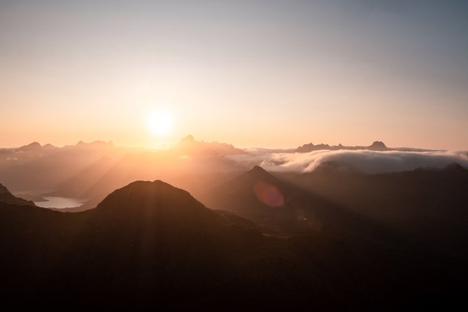 A breathtaking mountain landscape at sunrise or sunset, with golden sunlight streaming over jagged peaks above a sea of clouds and a small alpine lake visible below.