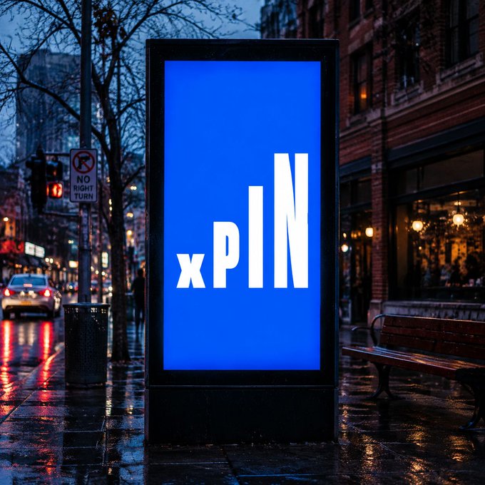 An outdoor advertising billboard displays 'xPIN' in large white text on a blue background, situated on a wet city street at dusk.