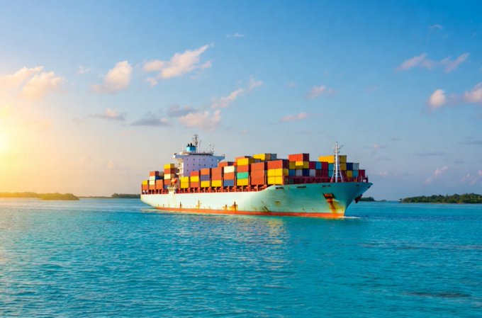 A large cargo ship loaded with colorful shipping containers sailing through turquoise ocean waters under a bright sunset sky.