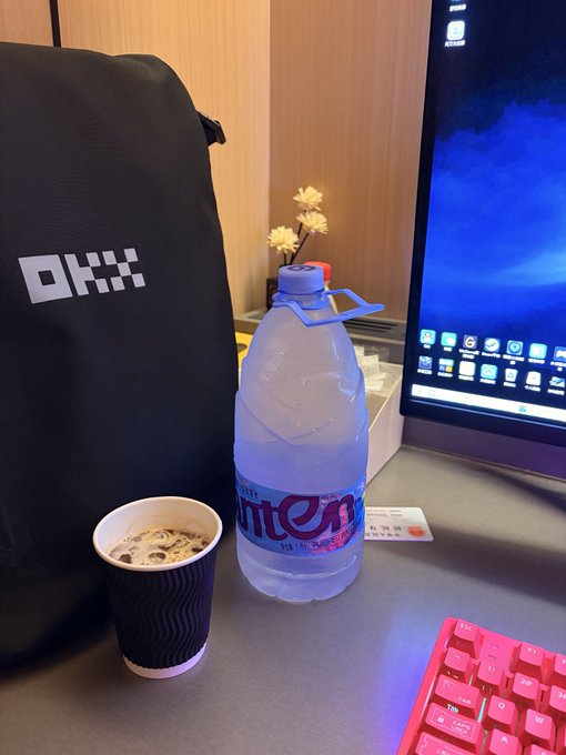 A black "OKX" branded bag sits beside a large bottle of "hint" water and a paper coffee cup on a desk with a pink mechanical keyboard. The setup reflects the caption's emphasis on bringing an oversized water bottle to fuel a long online session.