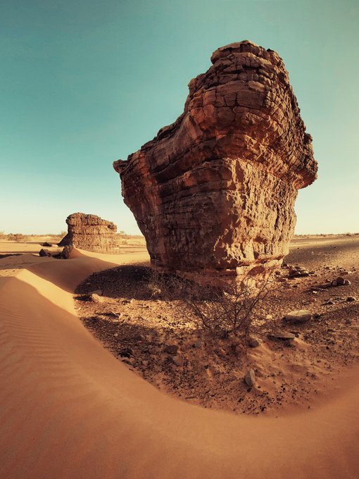 A large, balanced rock formation stands in a sandy desert landscape under a clear sky.