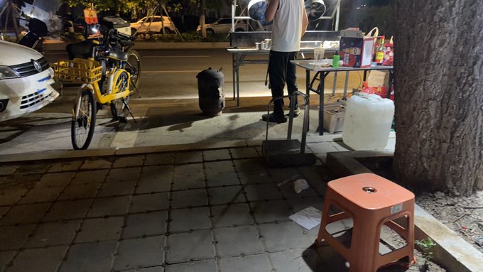 A lone yellow shared bike parked on the sidewalk at night near a bustling street-side barbecue stall where a vendor stands at his table. The image illustrates the post's humorous regret—riding the "little yellow bike" (a 3-yuan shared bike) to get late-night skewers, only to feel like the calories burned pedaling barely justified the indulgence compared to just driving there.