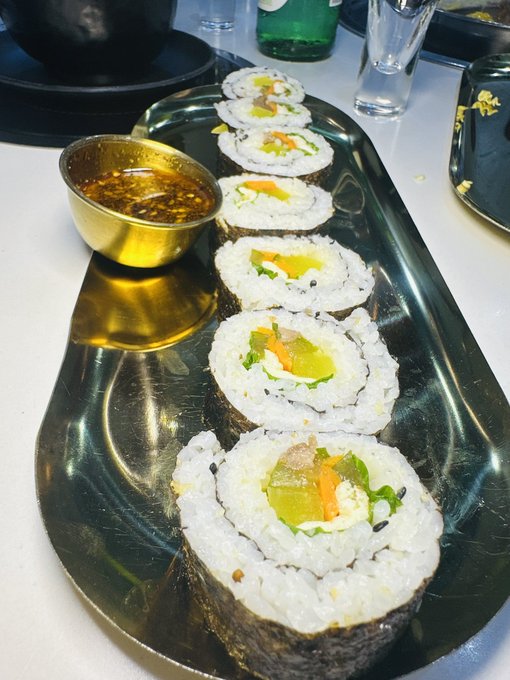 A sushi platter featuring six vegetable maki rolls (with carrots, cucumber, and greens) arranged in a row on a black tray alongside a gold dish of dipping sauce, fitting a casual "sunday dump" share of a leisurely meal.