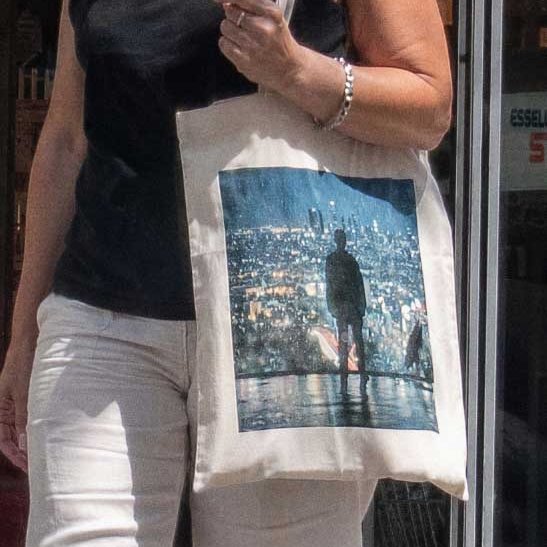 A person wearing a black top and white pants holds a beige tote bag featuring a printed image of a silhouetted figure overlooking a cityscape. They wear a silver bracelet on their wrist.