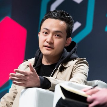 A man with dark hair, wearing a tan jacket over a black hoodie, gestures while seated during what appears to be a panel discussion or interview, with a vibrant pink and teal geometric backdrop behind him. The image likely features Justin Sun, the founder of TRON, discussing the network's new quantum-resistant signature feature now live on testnet.