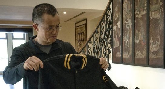 A man with glasses is examining a black and yellow letterman-style jacket indoors, with framed artwork visible on the wall behind him.