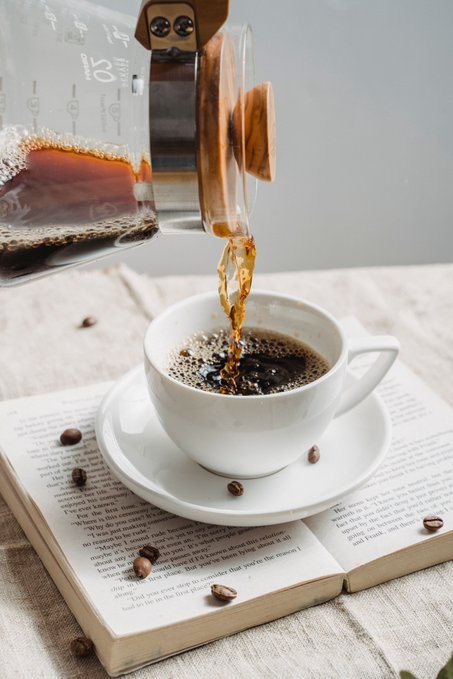 Coffee being poured from a glass measuring cup into a white cup resting on an open book, with coffee beans scattered on the pages.