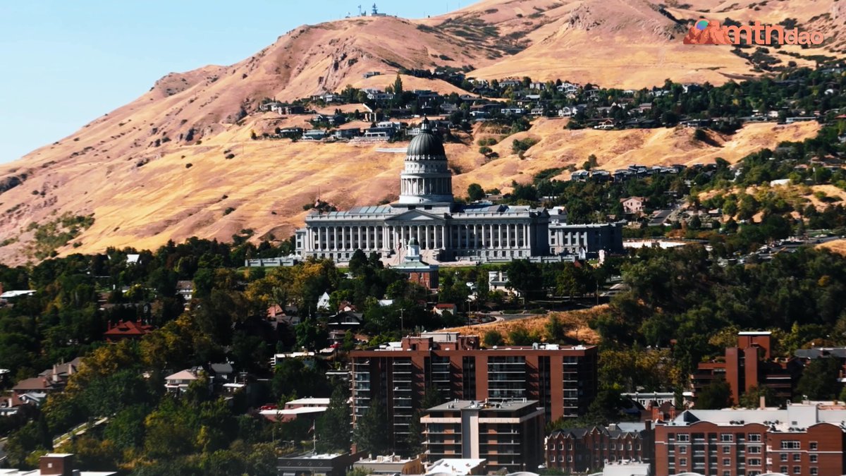 A scenic view of the Utah State Capitol building, featuring its prominent dome and classical architecture, set against a backdrop of golden-brown mountains with residential neighborhoods built into the hillside. A 'Kenhdao' watermark appears in the upper right corner.