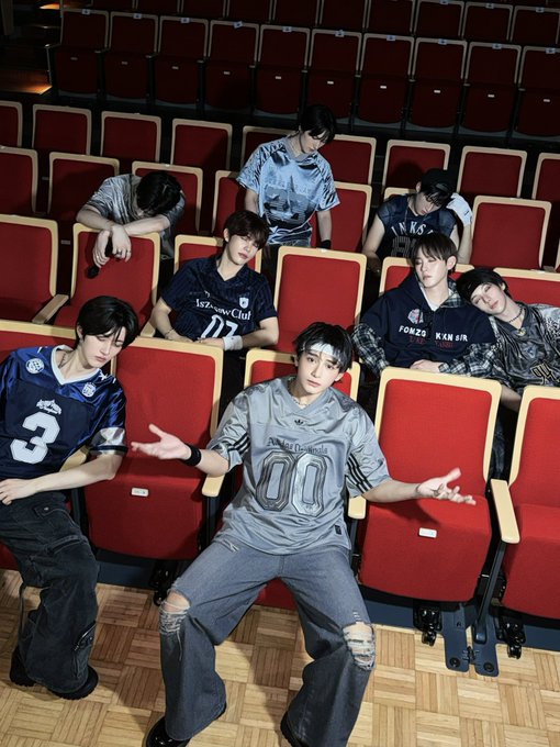 Seven young men in athletic-style jerseys (including numbers "3" and "00", "Izu...ow City", and "FORBES") pose confidently in red theater seats, promoting their group XODIAC ahead of the XBLISS FanCon event in Indonesia.