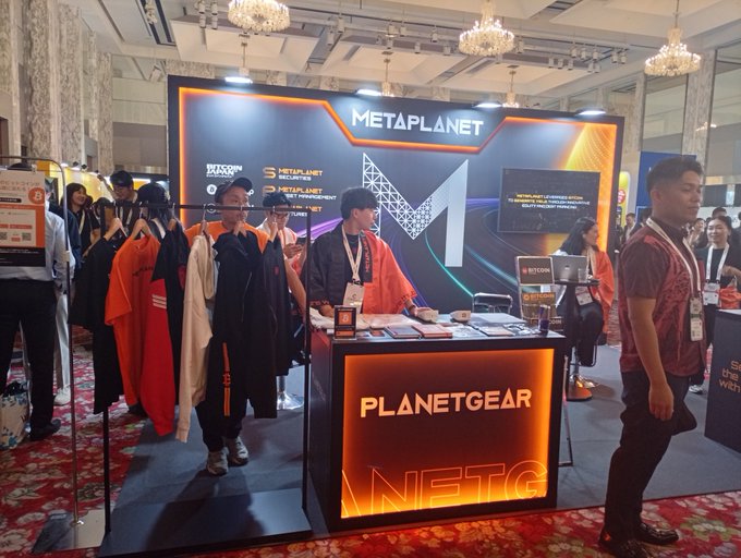 The image shows a Metaplanet booth at the WebX2026 conference in Tokyo, featuring branded displays with "PLANETGEAR" illuminated signage, clothing merchandise on racks, and promotional materials being staffed by representatives. The setup relates to the post by showcasing Metaplanet's active presence and engagement at a major crypto/Web3 event, highlighting the company's branding and promotional efforts that could support its growth narrative alongside Bitcoin's potential rally.
