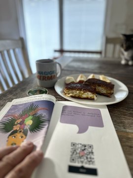 A first-person view of someone reading a book about "So Dead, So Alive" at a table, accompanied by a "MAGNOLIA BAKERY" mug and a plate with a pastry, while a cat sits in the background.