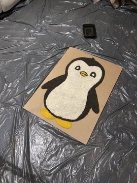 A small painting of a black and white penguin on a square canvas, resting on a wrinkled black plastic sheet next to a small black paint container.