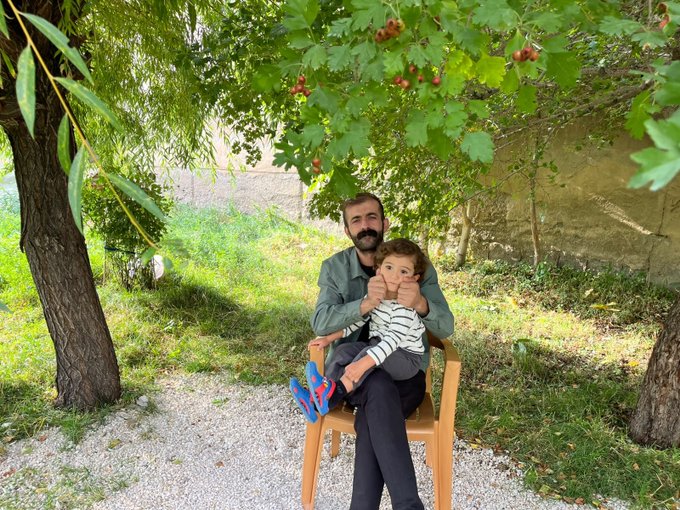A man with a beard sits on a chair outdoors under a tree with red berries, holding a young child on his lap. The child is wearing colorful sneakers and appears playful.