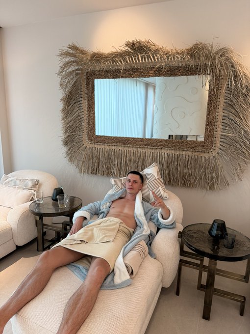 A young man reclining on a white chaise lounge in a living room, wearing an open robe, positioned beneath a large decorative straw-framed mirror with side tables holding lamps.
