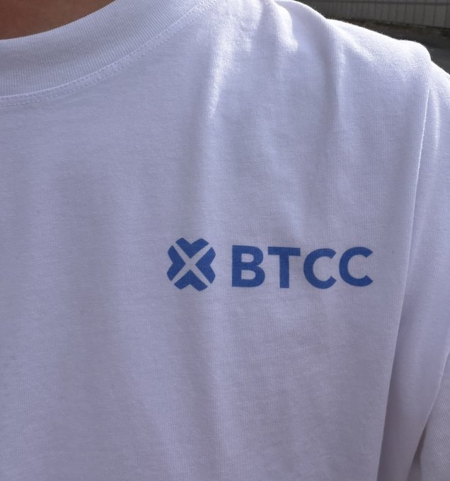 A close-up of a person wearing a white t-shirt featuring the blue "BTCC" logo (a stylized X emblem and text), shown as a promotional or branded shirt being worn casually outdoors.