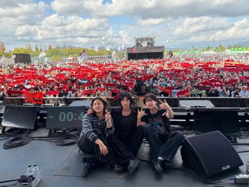 Three young people sit on the edge of a stage, making peace signs, with a massive crowd waving red glow sticks behind them at an outdoor concert.