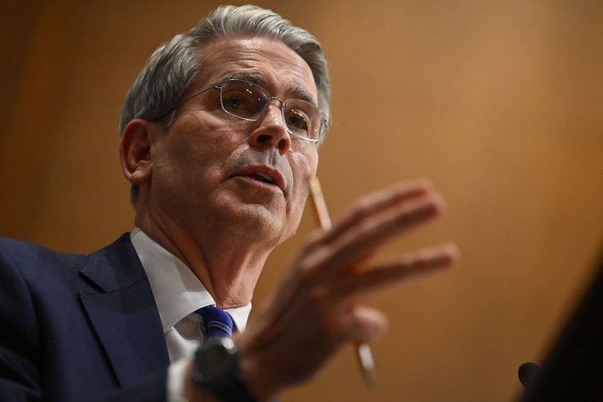 A man with glasses and gray hair in a dark suit and blue tie speaks while gesturing with his hand, with a pencil visible behind his ear.
