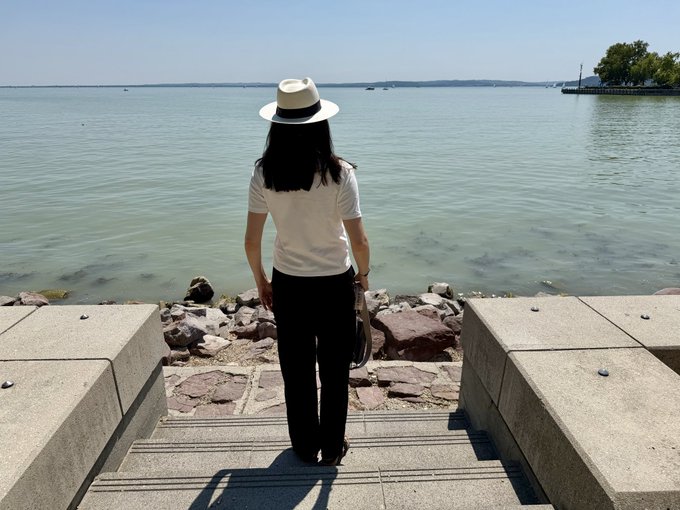 A woman in a white shirt, black pants, and a straw hat stands at the edge of a calm lake, gazing out at the water as if reflecting on her journey and motivation. The serene setting visually reinforces the post's message about remembering one's original purpose and staying grounded in personal belief rather than external validation.