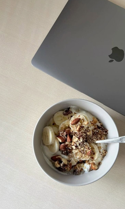 An overhead shot of a white bowl containing a yogurt parfait topped with banana slices, nuts, and seeds, sitting next to a closed Apple MacBook laptop on a light wooden desk.