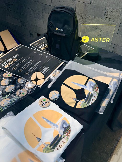 A merchandise display for 'ASTER' brand featuring a black backpack, t-shirts with graphic designs, badges, and an illuminated sign on a stone-textured surface.
