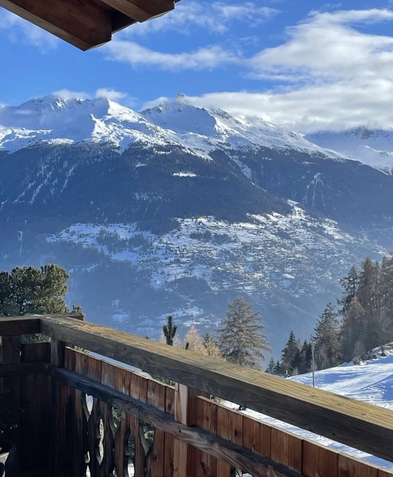 A wooden balcony overlooks a snow-covered Swiss alpine valley with scattered villages and evergreen trees, framing majestic snow-capped peaks under a partly cloudy sky. The serene mountain vista perfectly complements the post's dramatic tale of losing a crypto seed phrase to an Evian spring in the Swiss Alps.