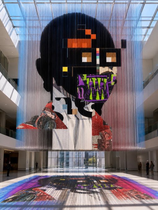 A large suspended art installation in a modern atrium depicts a pixelated portrait of a person wearing sunglasses, made of thousands of hanging strings with colorful elements.