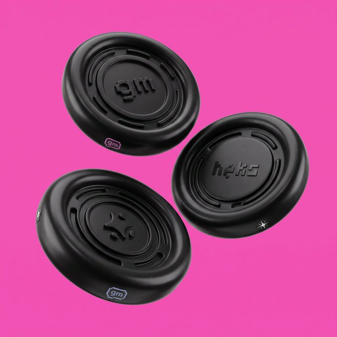 Three black disc-shaped objects with the engraved logos 'gm', 'heks', and 'gg' float against a bright pink background, with small 'gm' tags on the side.