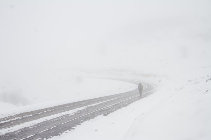 A snowy, foggy landscape with a dark curving road cutting through white hills, with a lone person walking near the bend.
