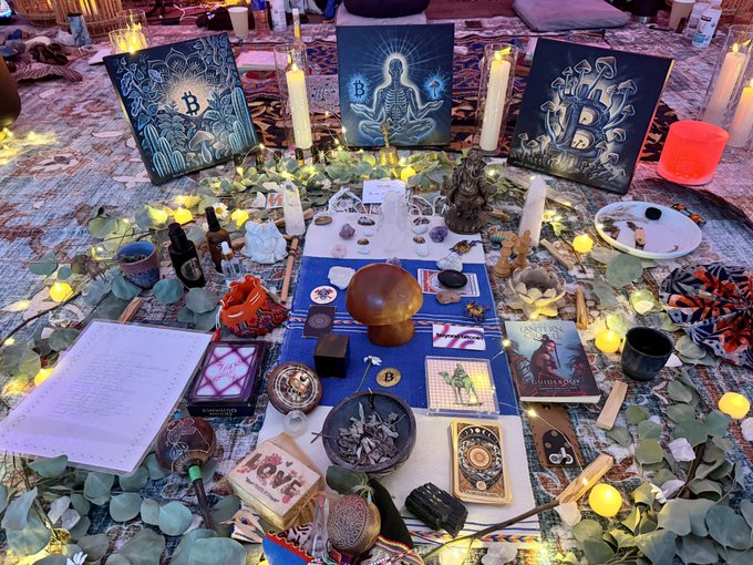 A spiritual altar with candles, tarot cards, crystals, eucalyptus leaves, and three paintings featuring Bitcoin symbols and a meditating Buddha-like figure.