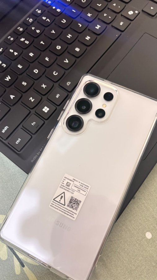 A white Samsung smartphone with a rear sticker bearing a QR code and identifier SAMSUNG ID: 3C69EB8A rests face-down on a black laptop keyboard, highlighting its multi-lens camera module.