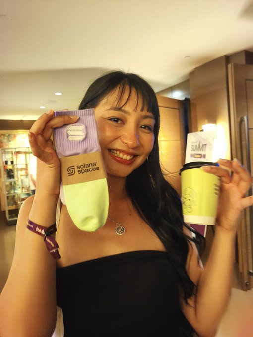 A smiling woman holds up two socks branded with "solana spaces" and "SUMMIT" logos in front of a mirror.