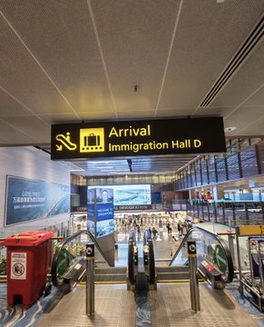 A glowing yellow "Arrival Immigration Hall D" sign hangs above escalators in an airport terminal, marking a traveler's arrival in the "Lion City." The scene captures the moment of stepping into Singapore, perfectly matching the post's announcement of arrival and the upcoming meetups with teachers and friends.