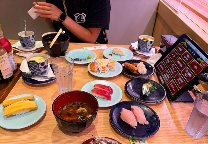 A conveyor belt sushi meal is laid out on a wooden table with various nigiri, miso soup, tamagoyaki, and a tablet menu visible.