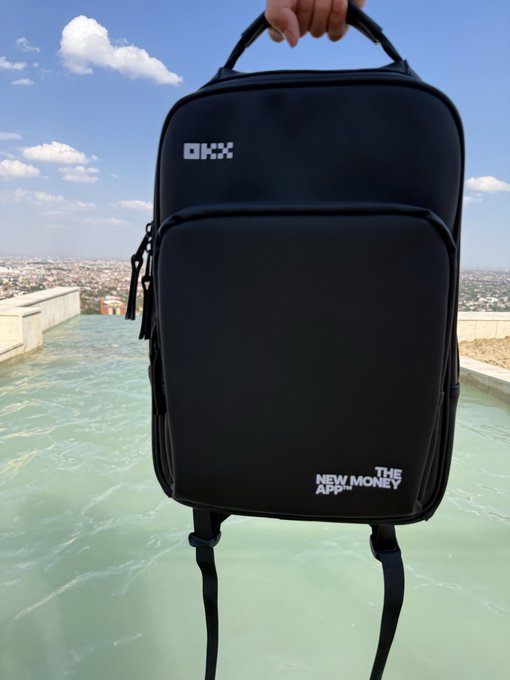 A black OKX branded backpack held up against a scenic rooftop with 'OKX' and 'THE NEW MONEY APP' text visible, set against a sky and cityscape background.