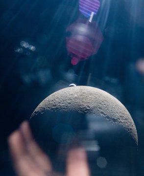 A detailed gray model of the moon hangs suspended against a dark blue background, with blurred hands and a small red figure visible behind it. The image visually echoes the caption's lunar ambition, turning the dream of going to the moon into a tangible, almost attainable scene.
