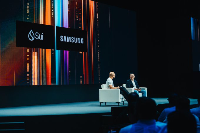 Two executives sit onstage beneath large screens displaying the Sui and Samsung logos, discussing their blockchain and mobile-technology partnership. The image supports the post’s claim that Sui and Samsung are collaborating on broader products aimed at helping people worldwide use Sui for everyday money transfers.