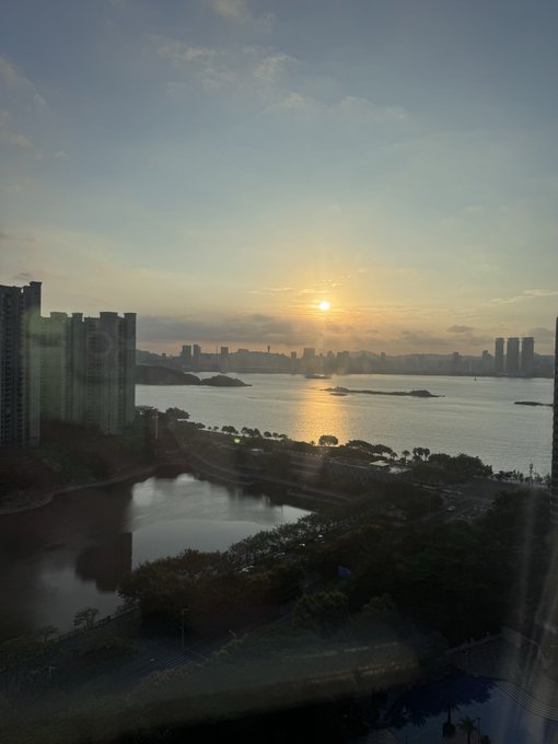 A scenic sunset view over a coastal city skyline shows the sun low on the horizon reflecting across the water, with high-rise buildings silhouetted in the distance.